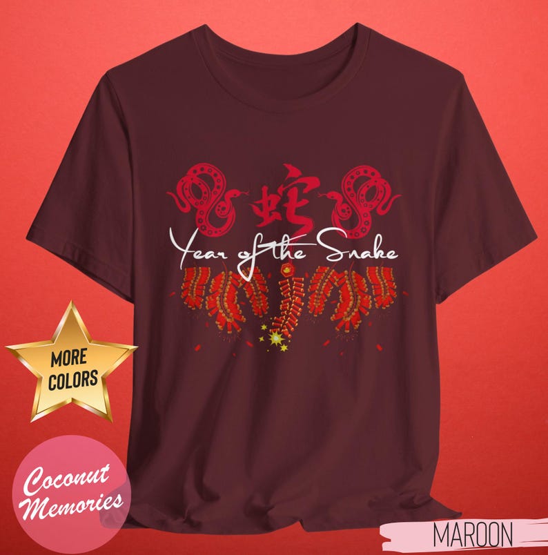 以下が含まれることがあります： 蛇と蛇の漢字が描かれた赤と白のデザインのマルーンのTシャツ。デザインの下に白い文字で「Year of the Snake」と印刷されています。