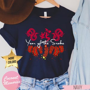 以下が含まれることがあります： 赤い中国語の文字と白い「蛇年」の文字が入ったネイビーブルーのTシャツ。デザインには、赤と白の花火と2匹の赤い蛇も含まれています。