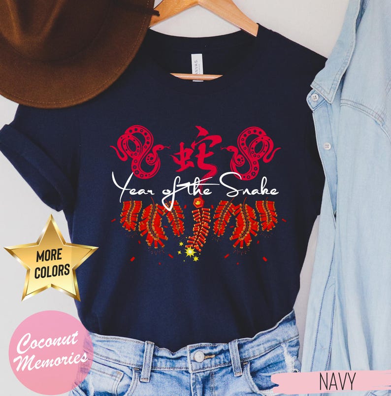 以下が含まれることがあります： 赤い中国語の文字と白い「蛇年」の文字が入ったネイビーブルーのTシャツ。デザインには、赤と白の花火と2匹の赤い蛇も含まれています。
