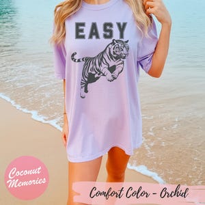 Pu&ograve; includere: Una maglietta oversize color lavanda con la parola "EASY" stampata in nero e una grafica di tigre in bianco e nero. La maglietta ha una vestibilit&agrave; rilassata e un collo rotondo.