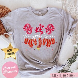 以下が含まれることがあります： 白い蛇と爆竹の白いグラフィックが描かれた灰色のTシャツ。グラフィックの下に「蛇年」の文字が印刷されています。