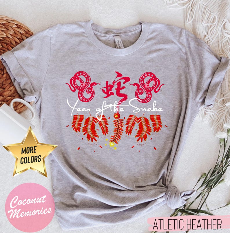 以下が含まれることがあります： 白い蛇と爆竹の白いグラフィックが描かれた灰色のTシャツ。グラフィックの下に「蛇年」の文字が印刷されています。