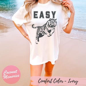 Pu&ograve; includere: Una maglietta bianca con la parola "EASY" stampata in nero sul davanti. Un grafico di tigre bianco e nero si trova sotto il testo. La maglietta &egrave; indossata da un modello in piedi su una spiaggia.