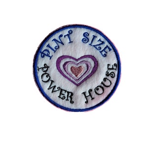 May include: A white embroidered patch with blue trim. The patch features a heart with pink and purple layers and the text "Pint Size Power House".