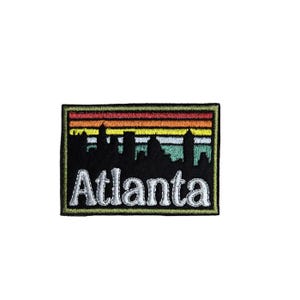 May include: A rectangular embroidered patch featuring a black silhouette of the Atlanta skyline against a horizontal striped background of red, orange, yellow, light blue, and teal. The word Atlanta is stitched in white thread at the bottom.
