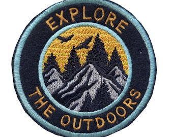 May include: A round, black and blue embroidered patch with the text "Explore the Outdoors" in yellow thread. The patch features a mountain range with trees and a yellow sun in the background.