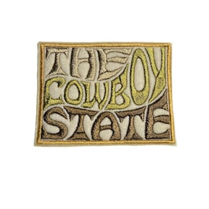 May include: Embroidered patch with the words "THE COWBOY STATE" in a stylized font. The patch has a rectangular shape with a gold border. The text is in shades of beige, brown, and gold.