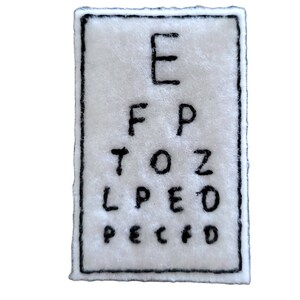 May include: A white felt patch embroidered with a black eye chart. The chart features the letters E, F, P, T, O, Z, L, P, E, D, and a black border.