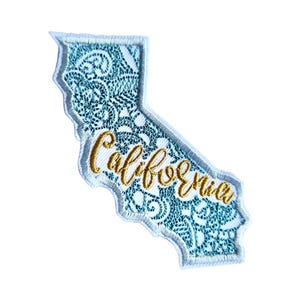 Puede incluir: Un contorno blanco del estado de California con un diseño estampado azul y blanco y la palabra "California" bordada en oro.