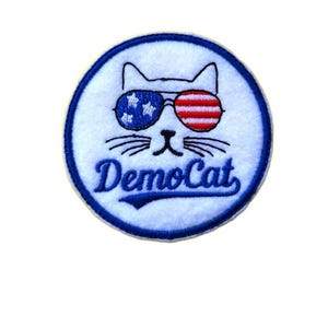 May include: A white cat wearing sunglasses with an American flag design. The cat's eyes are red, white, and blue. The text "DemoCat" is written in blue cursive letters below the cat's face. The image is a round, blue-bordered patch.