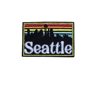 May include: Black and white embroidered patch with a rainbow sunset and a skyline of Seattle, Washington. The text "Seattle" is in white.