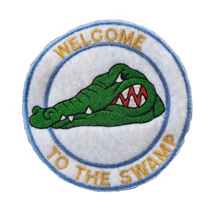May include: A white, round embroidered patch with a blue border. The patch features a green alligator with red teeth and the text "Welcome to the Swamp" in gold.