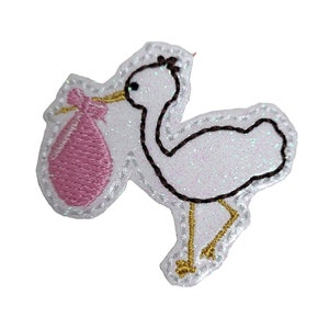 May include: A white glitter stork with brown and gold embroidery carrying a pink baby bag with a gold bow.