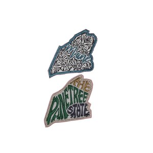 May include: Two embroidered patches shaped like the state of Maine. One patch is white with a blue outline and a black and white mandala design with the word "Maine" in blue. The other patch is pink with a green and gold outline and the words "The Pine Tree State" in green and gold.