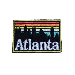 May include: An embroidered patch with a black silhouette of the Atlanta skyline against a colorful sunset background. The text "Atlanta" is embroidered in white below the skyline.