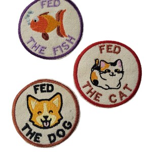 May include: Three embroidered patches with a white background and colorful embroidery. The patches are round and feature a fish, a cat, and a dog. The text on the patches reads "Fed the Fish", "Fed the Cat", and "Fed the Dog".