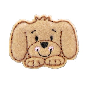 May include: A brown felt applique of a dog with pink cheeks and a black nose. The dog is smiling and has its paws crossed in front of it.