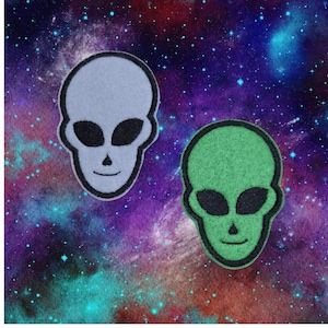 May include: Two felt alien head patches, one white and one green, with black eyes and a smiling mouth. The patches are set against a galaxy-themed background.