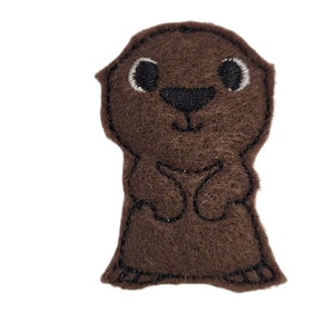 May include: A brown felt appliqué of a cartoon otter with black eyes, nose, and mouth. The otter is standing with its arms crossed.