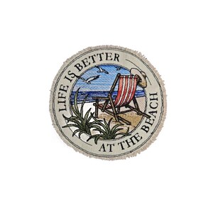 May include: A round, fringed fabric coaster featuring an embroidered beach scene with a red and white striped lounge chair, green plants, and seagulls. The text "LIFE IS BETTER AT THE BEACH" is embroidered in black around the circular border.