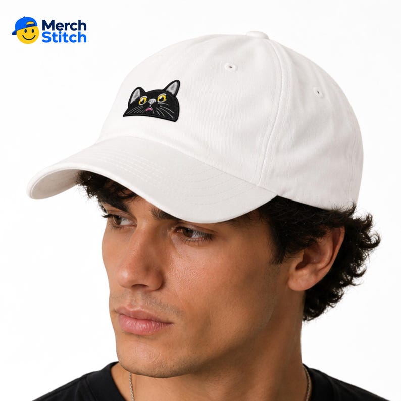 May include: A white baseball cap featuring an embroidered graphic of a black cat head with yellow eyes and white whiskers peeking out from the bottom edge. The cap has a curved brim and a structured crown with a button on the top center.