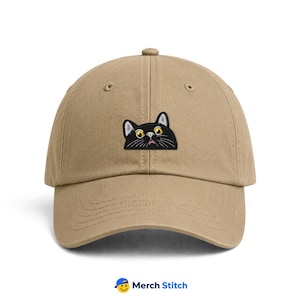 May include: A tan baseball cap featuring an embroidered patch of a black cat head with large yellow eyes and a small pink nose. The cat is depicted as if peeking over the center seam of the cap. The cap has a curved brim and a button on top.