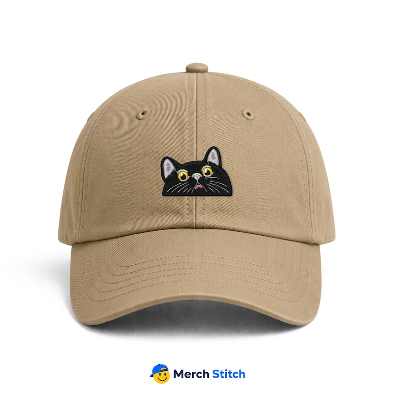 May include: A tan baseball cap featuring an embroidered patch of a black cat head with large yellow eyes and a small pink nose. The cat is depicted as if peeking over the center seam of the cap. The cap has a curved brim and a button on top.