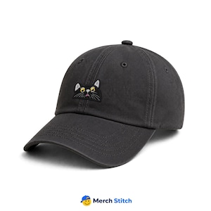 May include: A charcoal gray baseball cap featuring an embroidered patch of a black cat face with wide yellow eyes, white whiskers, and a small pink mouth. The cap has a curved brim, a button on top, and stitched eyelets for ventilation.