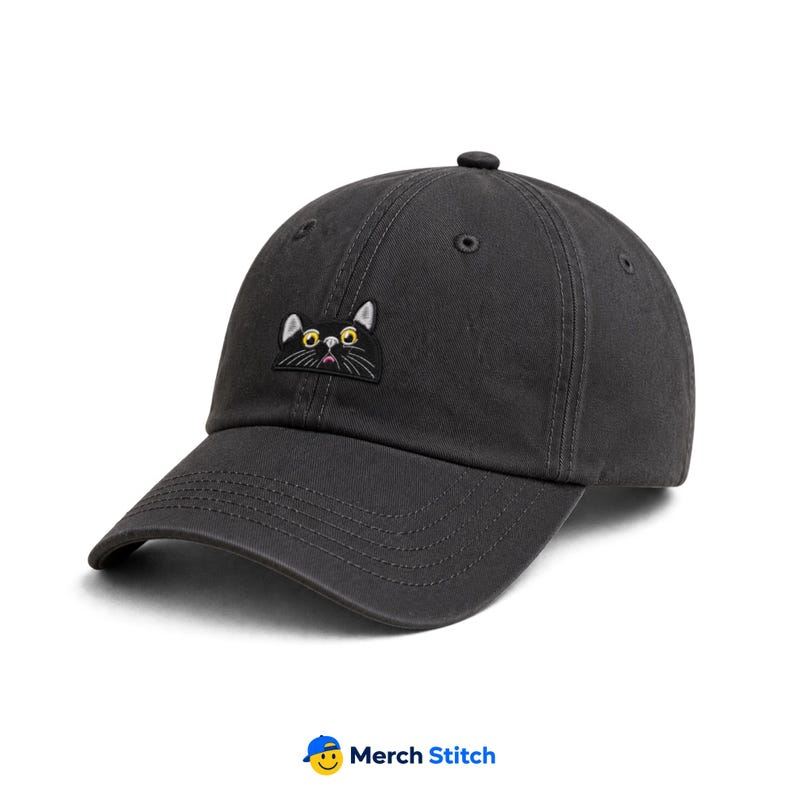 May include: A charcoal gray baseball cap featuring an embroidered patch of a black cat face with wide yellow eyes, white whiskers, and a small pink mouth. The cap has a curved brim, a button on top, and stitched eyelets for ventilation.