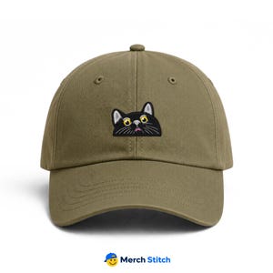 May include: An olive green baseball cap featuring an embroidered patch of a black cat face with large yellow eyes, white whiskers, and a small pink mouth. The cap has a structured crown, a curved brim, and stitched eyelets for ventilation.