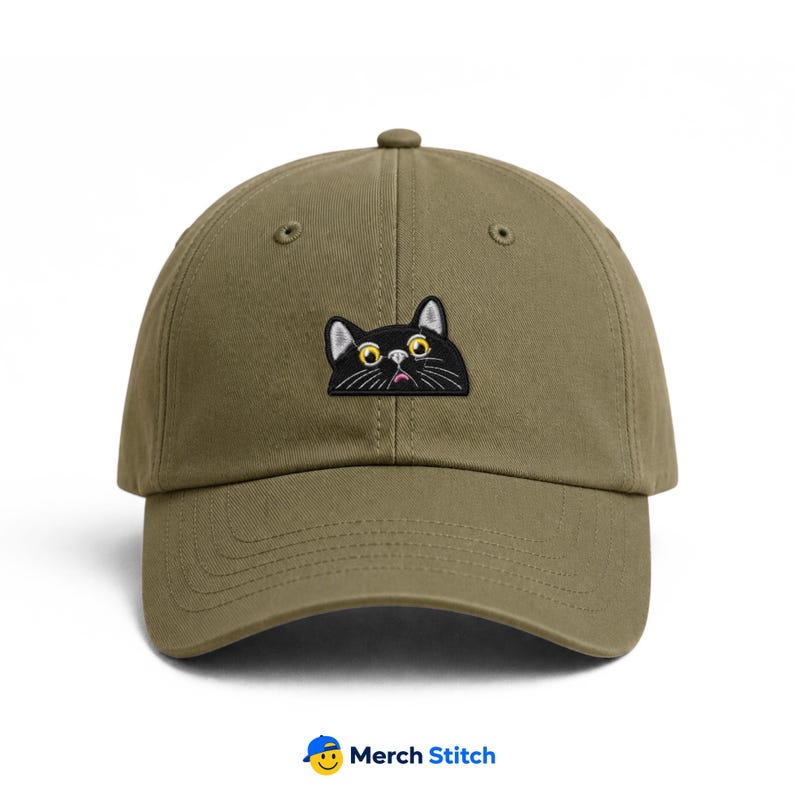 May include: An olive green baseball cap featuring an embroidered patch of a black cat face with large yellow eyes, white whiskers, and a small pink mouth. The cap has a structured crown, a curved brim, and stitched eyelets for ventilation.