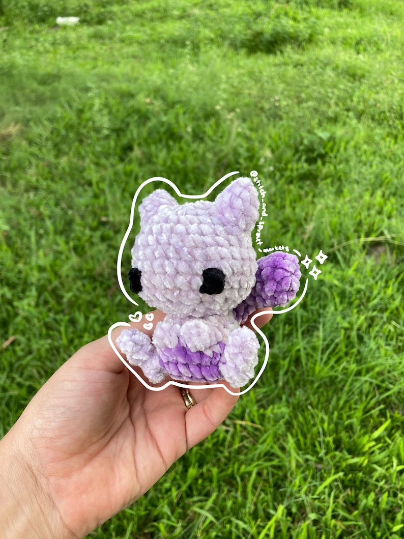 May include: A small, purple and white crocheted plush toy in the shape of a cat. The toy is being held in a person's hand.