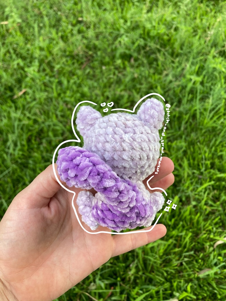 May include: A purple and white crocheted plush toy in the shape of a cartoon character. The toy is holding its tail in its hands. The toy is on a green background.