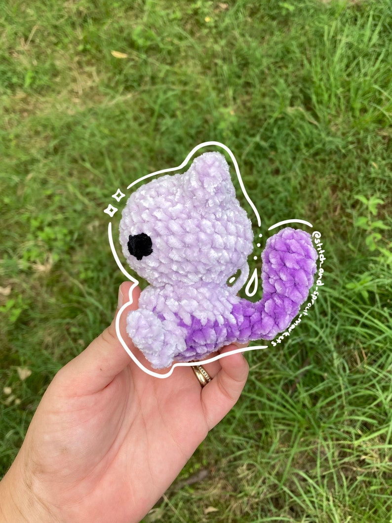 May include: A purple and white crocheted plush toy in the shape of a cat. The toy has a black eye and a white outline. The toy is being held in a hand.