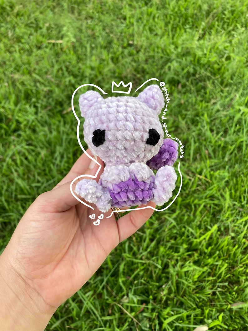 May include: A small, crocheted purple and lavender plush toy in the shape of a cat. The toy has black eyes and a crown on its head. The toy is being held in a hand against a green grassy background.