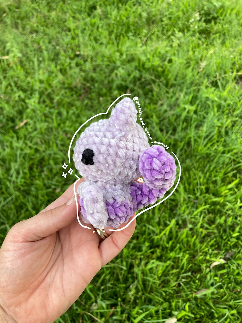 May include: A small, crocheted purple and lavender plush toy in the shape of an octopus. The toy has black eyes and is being held in a hand.