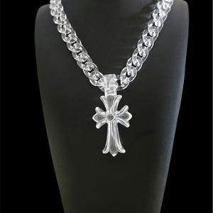 May include: A clear, transparent acrylic chain necklace featuring a large, ornate cross pendant. The pendant has a stylized, flared design with a central circular detail, suspended from a thick, interlocking link chain made of the same material.
