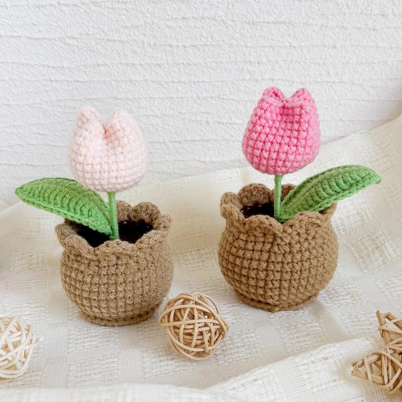 May include: Two crochet tulip flowers in brown pots. One tulip is light pink, the other is hot pink. Each flower has a green stem and leaf. A small woven ball sits between the pots. The pots have a scalloped edge.