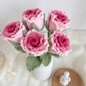 May include: A bouquet of five crocheted roses in shades of pink, with green crocheted leaves. The roses are arranged in a white vase.