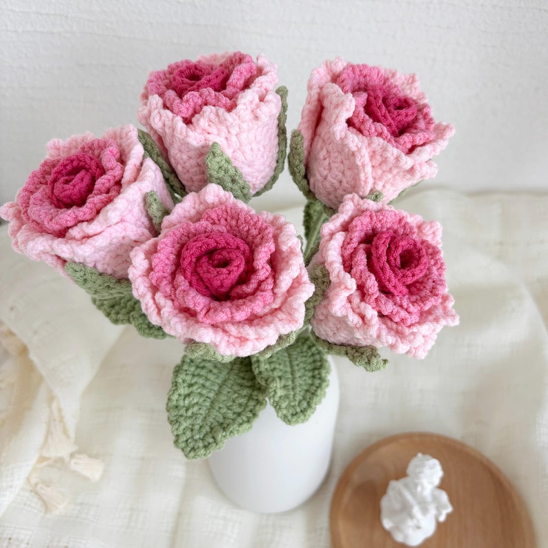 May include: A bouquet of five crocheted roses in shades of pink, with green crocheted leaves. The roses are arranged in a white vase.