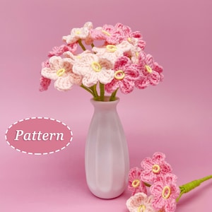 Puede incluir: Un ramo de flores de ganchillo rosa y blanco en un jarrón blanco. Las flores están dispuestas en un jarrón con una superficie texturizada. El jarrón está sobre una superficie rosa. El texto "Pattern" está escrito en un óvalo blanco con un borde discontinuo.