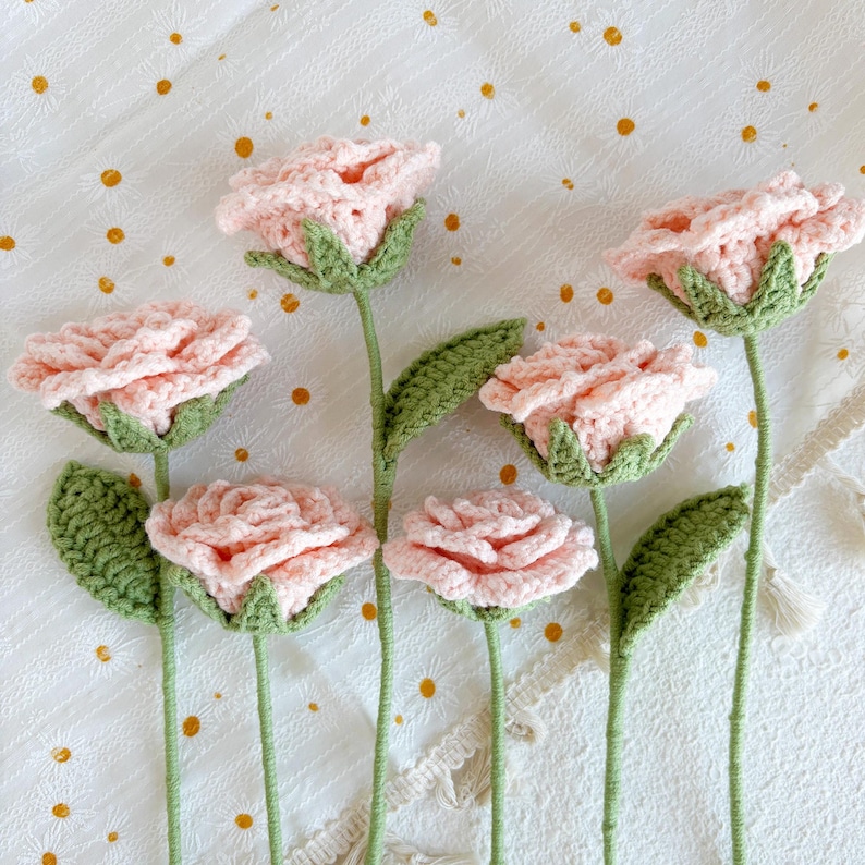 May include: Six crocheted pink roses with green stems and leaves on a white background with gold polka dots.
