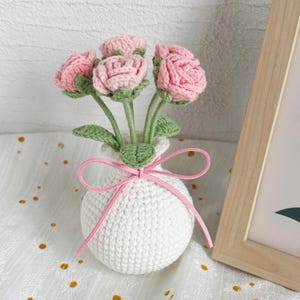 May include: A handmade crochet flower arrangement featuring pink roses in a white vase. The vase is tied with a pink ribbon. The flowers have green stems and leaves, creating a charming, handcrafted floral display.