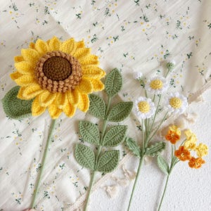 May include: Three crocheted flower stems with green leaves. The first stem has a yellow crocheted sunflower with a brown center. The second stem has white crocheted daisies. The third stem has orange crocheted flowers.