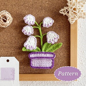 May include: A handcrafted crochet flower arrangement in a purple pot. The white flowers have purple edges, green leaves, and a green stem. The arrangement is on a corkboard with a wooden frame. A tag and a label with the word "Pattern" are also visible.