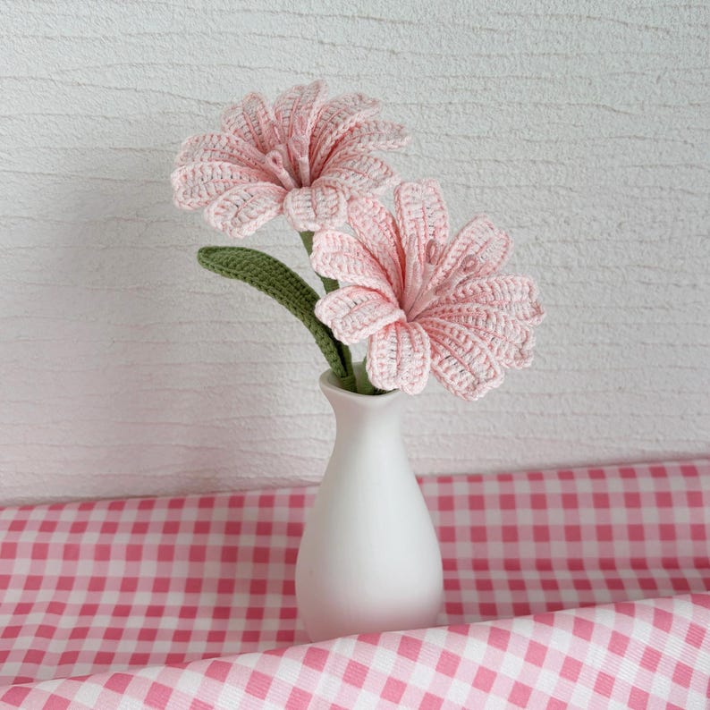 May include: Two pink crocheted flowers in a white vase on a pink and white gingham tablecloth.