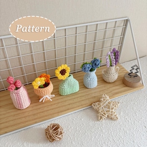 Puede incluir: Cinco pequeños arreglos florales de ganchillo en jarrones de varios colores pastel, incluyendo rosa, naranja, verde menta, azul claro y crema. El conjunto presenta diversos diseños florales sobre un estante de madera con rejilla blanca.