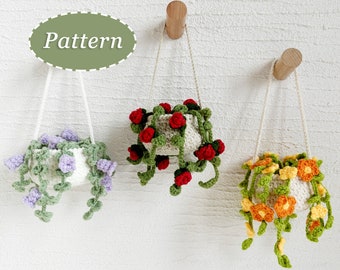 Op de afbeelding: Drie handgemaakte gehaakte hangende plantenbakken met bloemmotieven. De plantenbakken zijn wit met paarse, rode en oranje bloemen en groene wijnstokken. De plantenbakken hangen aan houten pinnen aan een witte muur. Het woord "Pattern" staat in een groene ovaal.