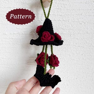 May include: A black crochet witch hat with red roses and a bat, hanging from a green cord. The word "Pattern" is in a maroon oval. The hat is decorated with red roses.