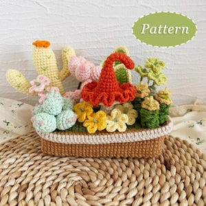 May include: A crocheted miniature garden scene featuring a variety of colorful plants and flowers, including a cactus, mushrooms, and other floral elements. The scene is contained within a brown basket with a cream-colored trim. The word "Pattern" is in a green oval.